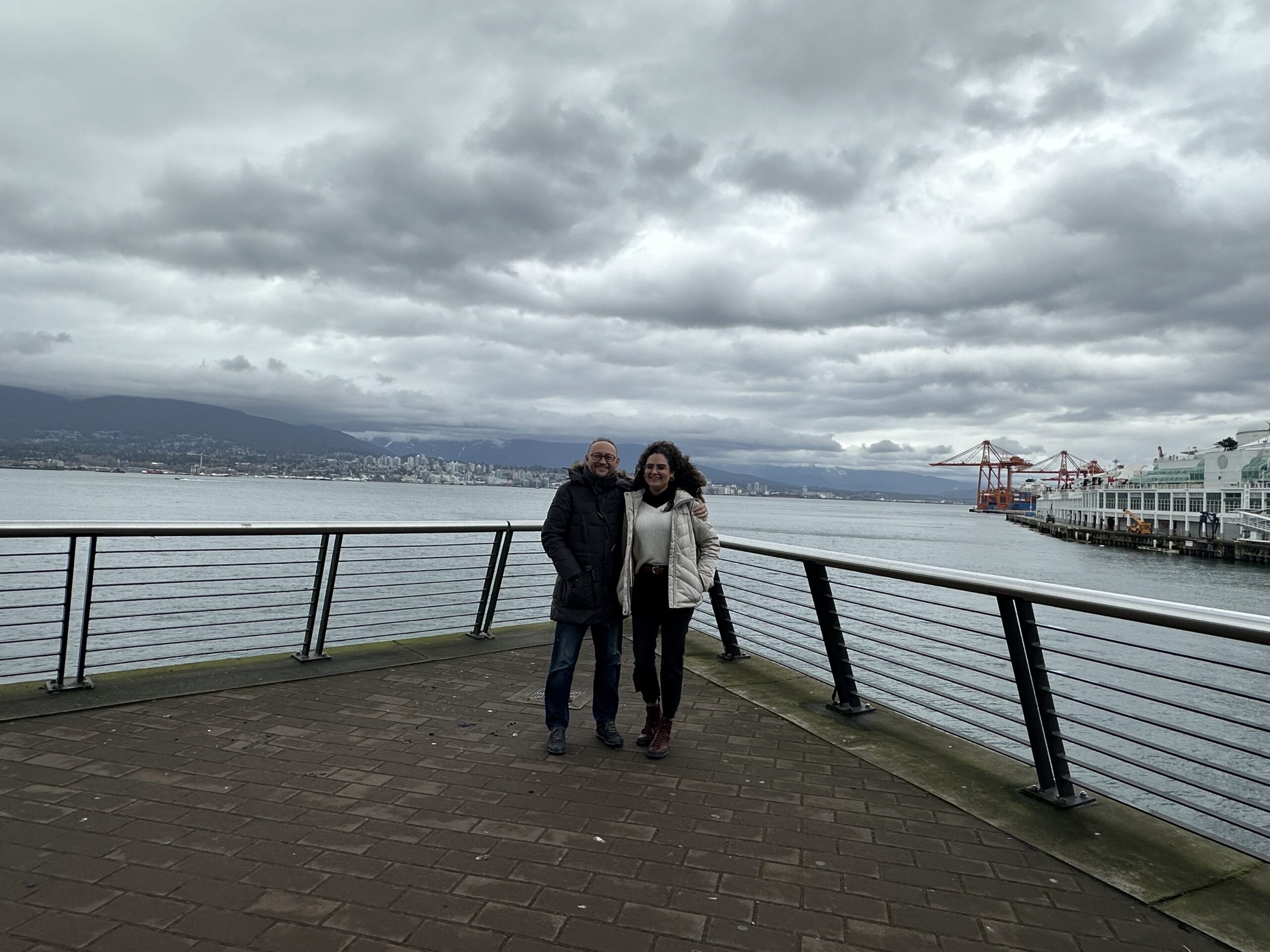 A couple posing on the Canada Place pier in Vancouver, with the ocean and harbor cranes in the background.