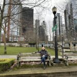 A man in a jacket sitting on a wooden bench at Harbour Green Park, Vancouver, with downtown skyscrapers and bare trees in the background on a cloudy day.