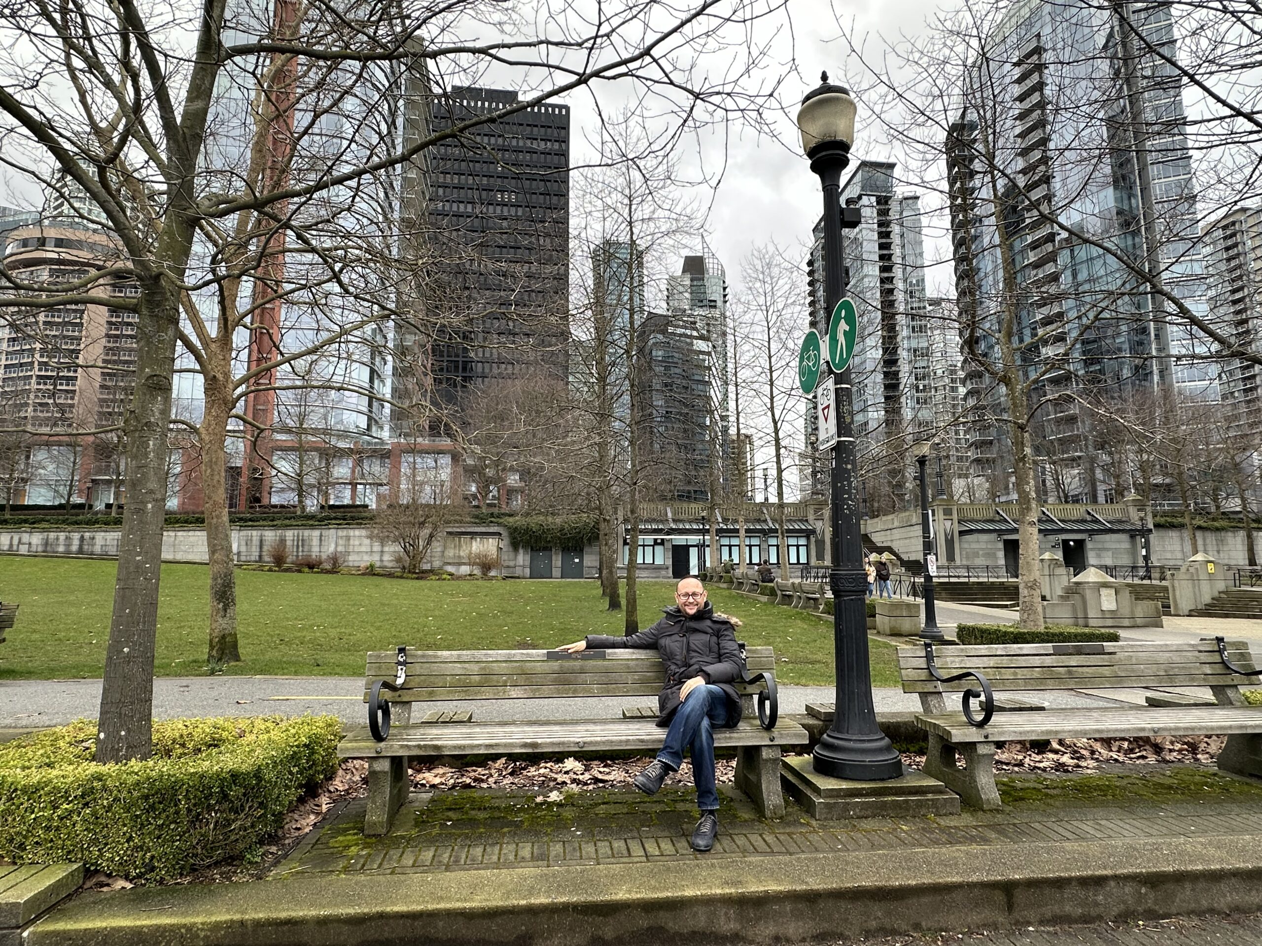 A man in a jacket sitting on a wooden bench at Harbour Green Park, Vancouver, with downtown skyscrapers and bare trees in the background on a cloudy day.