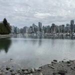 Vancouver Stanley Park Seawall'dan (deniz duvarı yürüyüş yolu) sakin iç limana doğru çekilmiş, karşı kıyıdaki şehir merkezinin silüetini gösteren geniş bir panoramik manzara. Ön planda çakıllı bir sahil, solda yaprak dökmeyen ağaçlar ve ortada bulutlu bir gökyüzü altında beyaz teknelerle dolu bir marina görülüyor.