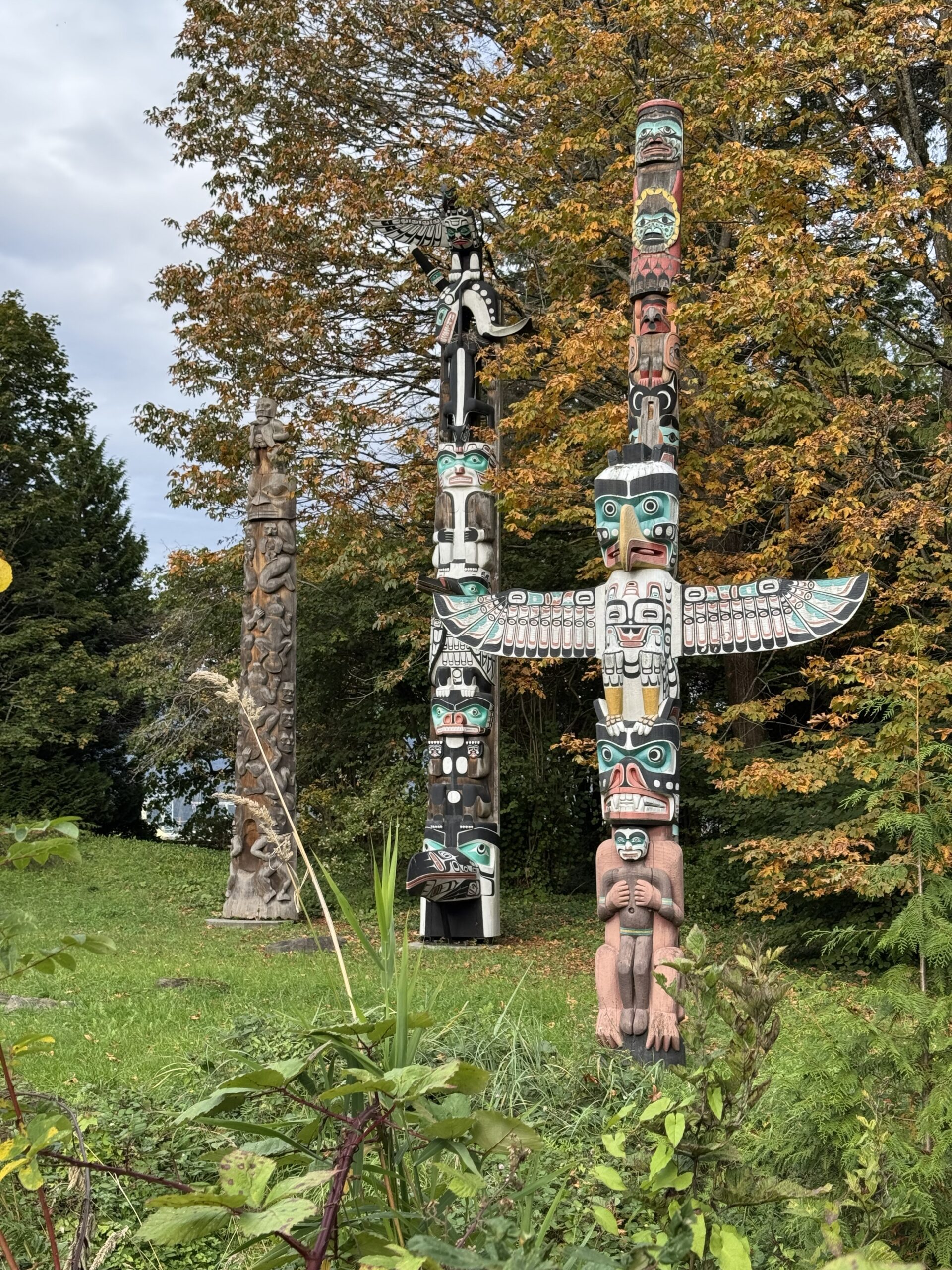 Three large, brightly painted Indigenous totem poles standing tall on a grassy slope at Stanley Park, Vancouver, BC. They are surrounded by diverse trees with green and autumn-orange leaves under a cloudy sky. The main pole in the foreground features a Thunderbird with outstretched wings.