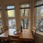 A cozy, sunlit dining area within a guest suite at Times Square Suites Hotel in Vancouver's West End, featuring a wooden table with four chairs by a large four-panel bay window overlooking tree-lined city streets.