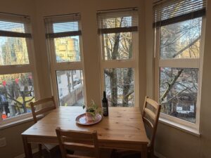 A cozy, sunlit dining area within a guest suite at Times Square Suites Hotel in Vancouver's West End, featuring a wooden table with four chairs by a large four-panel bay window overlooking tree-lined city streets.