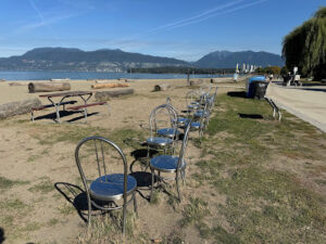 A row of empty metal chairs on the sand at Kitsilano Beach, Vancouver, overlooking the ocean and the North Shore Mountains.