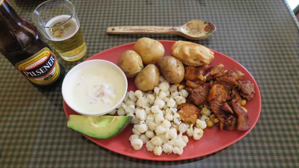 A traditional Ecuadorian dish called Carnes Coloradas served on a red plate in Cotacachi, featuring marinated pork, boiled potatoes, mote (hominy), avocado, an empanada, and a side of white sauce, accompanied by a bottle of Pilsener beer.
