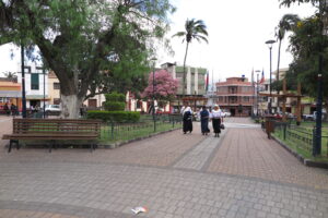 A wide-angle view of a peaceful public plaza in Cotacachi, Ecuador, featuring a paved walkway, a wooden bench under a large tree, and indigenous women in traditional clothing walking through the square.