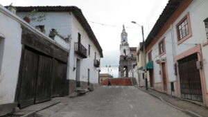 A cobblestone street in Cotacachi, Ecuador, looking towards a historic church tower with a red-tiled top, flanked by traditional white-walled colonial buildings with wooden balconies.