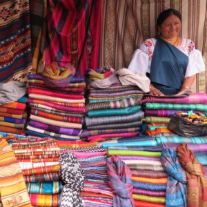 A vibrant stall at the Otavalo Market in Ecuador, showcasing stacks of colorful handwoven Andean textiles, blankets, and scarves, with an indigenous woman in traditional dress standing behind the counter.