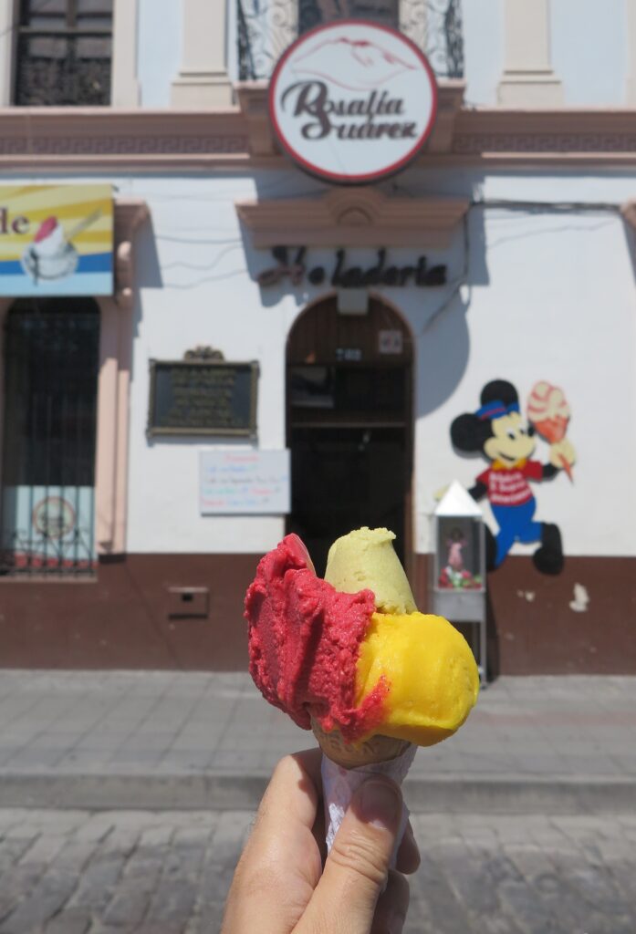 A person holding a colorful three-scoop fruit ice cream cone in front of the historic Rosalía Suárez ice cream shop in Ibarra, Ecuador, with the shop's logo and a Mickey Mouse mural in the background.