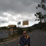 A travel blogger taking a selfie on a paved mountain road in Ecuador, with signs pointing toward Laguna de Cuicocha under a dramatic cloudy sky.
