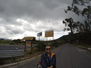 A travel blogger taking a selfie on a paved mountain road in Ecuador, with signs pointing toward Laguna de Cuicocha under a dramatic cloudy sky.