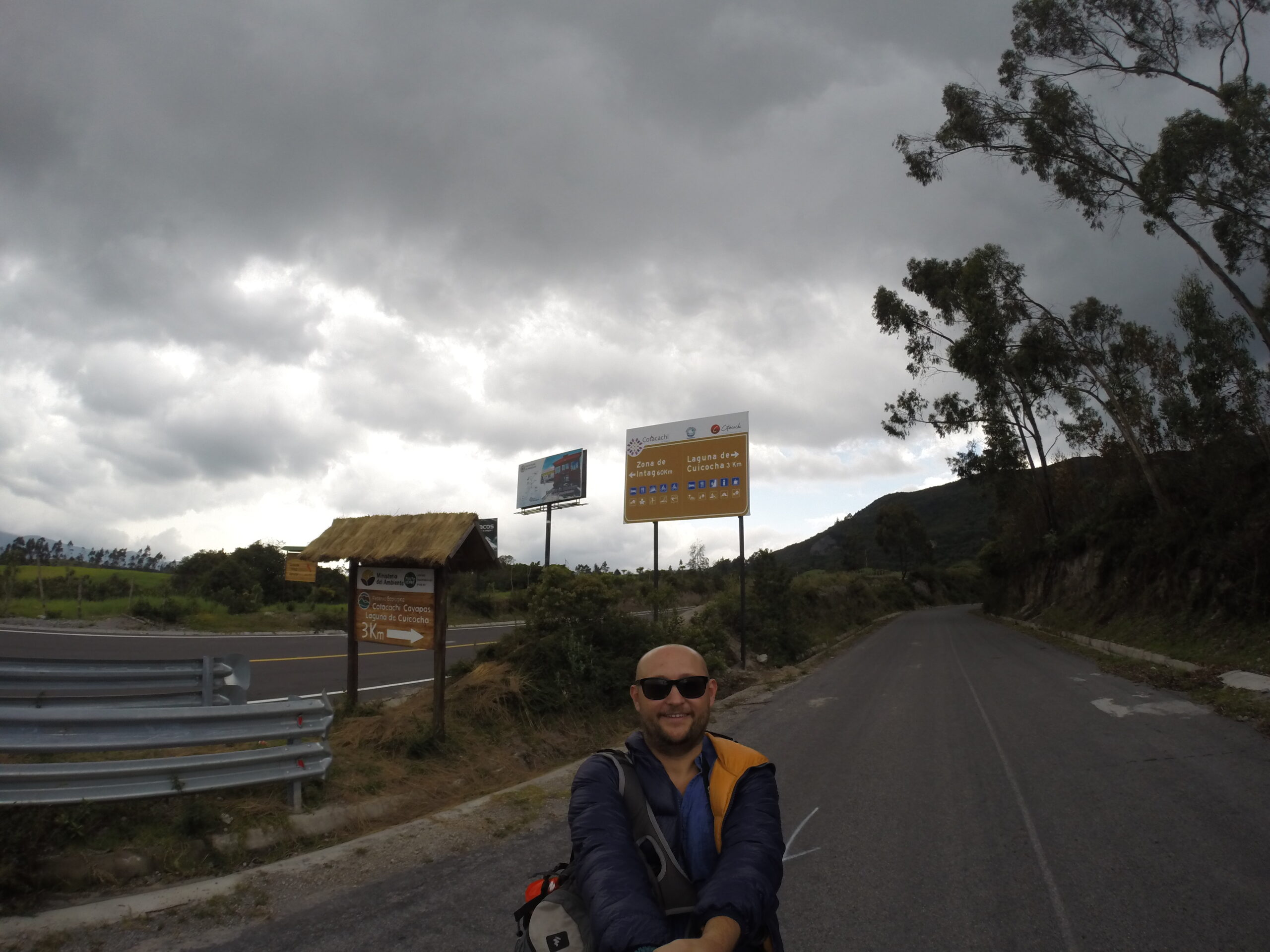 A travel blogger taking a selfie on a paved mountain road in Ecuador, with signs pointing toward Laguna de Cuicocha under a dramatic cloudy sky.