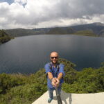 A travel blogger taking a high-angle selfie with a selfie stick at a viewpoint overlooking the vast Laguna de Cuicocha crater lake in Ecuador, featuring the two distinct islands in the center and lush green caldera walls under a cloudy sky.