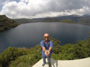 A travel blogger taking a high-angle selfie with a selfie stick at a viewpoint overlooking the vast Laguna de Cuicocha crater lake in Ecuador, featuring the two distinct islands in the center and lush green caldera walls under a cloudy sky.