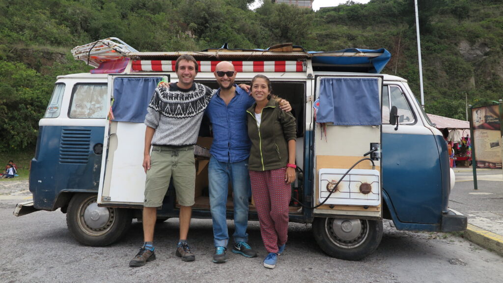 A travel blogger standing with an Argentine couple in front of their blue and white Volkswagen camper van at Laguna de Cuicocha, Ecuador, with the "Kombiando por America" project visible.