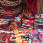 A vibrant stall at the Otavalo Market in Ecuador, showcasing stacks of colorful handwoven Andean textiles, blankets, and scarves, with an indigenous woman in traditional dress standing behind the counter.