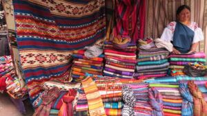 A vibrant stall at the Otavalo Market in Ecuador, showcasing stacks of colorful handwoven Andean textiles, blankets, and scarves, with an indigenous woman in traditional dress standing behind the counter.