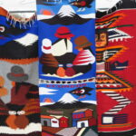 Close-up of colorful handwoven wall hangings at the Otavalo Market in Ecuador, featuring traditional patterns of Andean people, Condors, Cotopaxi volcano, and geometric designs.