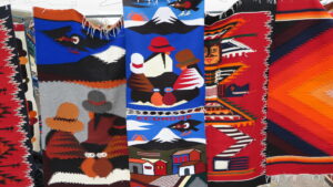 Close-up of colorful handwoven wall hangings at the Otavalo Market in Ecuador, featuring traditional patterns of Andean people, Condors, Cotopaxi volcano, and geometric designs.