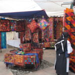 A vibrant stall at Otavalo Market in Ecuador filled with hand-embroidered pillows, floral textiles, and woven wall hangings featuring Andean portraits and landscapes, with local shoppers browsing.