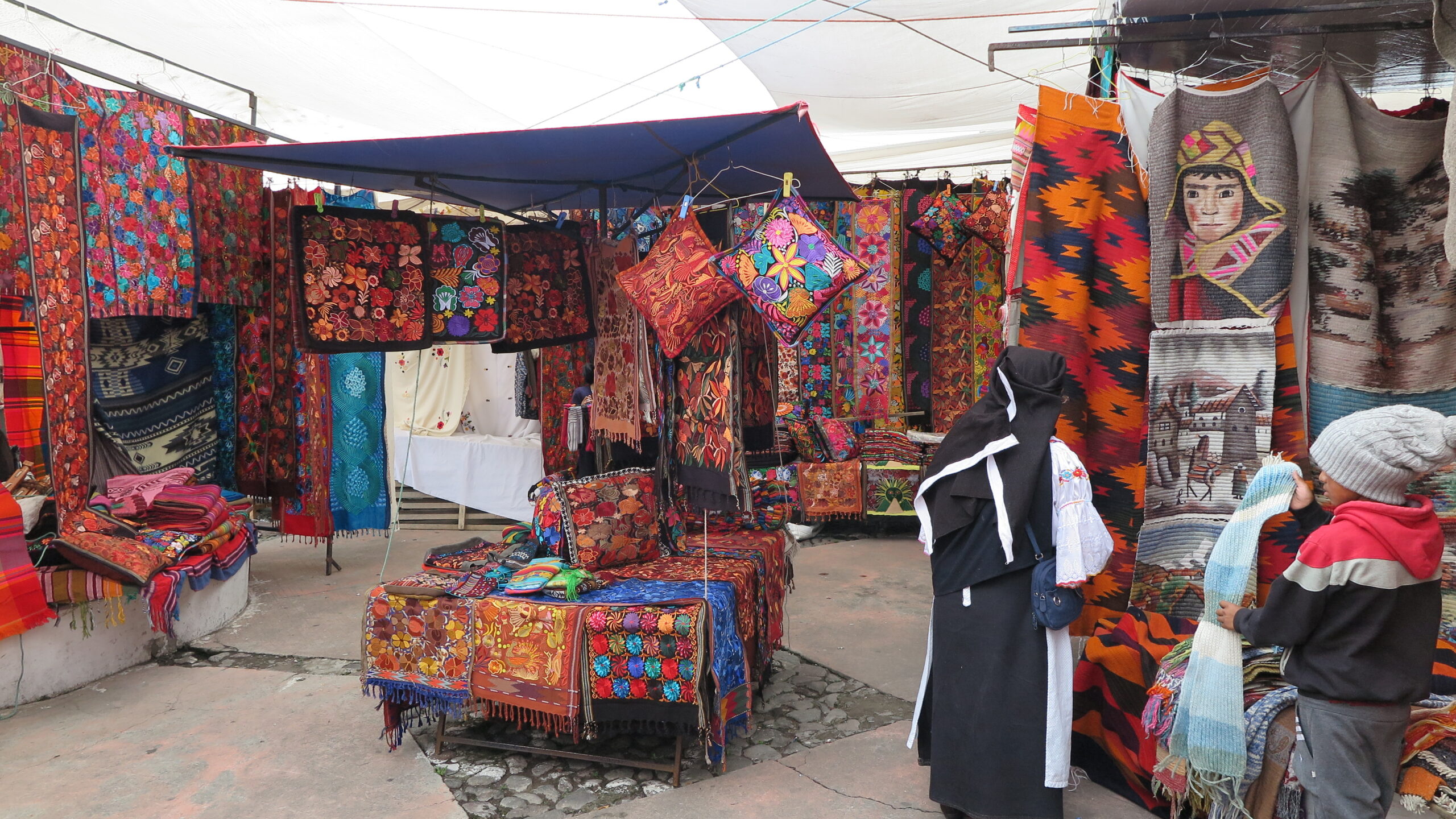 A vibrant stall at Otavalo Market in Ecuador filled with hand-embroidered pillows, floral textiles, and woven wall hangings featuring Andean portraits and landscapes, with local shoppers browsing.