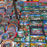 A vibrant display at Otavalo Market in Ecuador featuring hand-painted wooden bowls and Tigua-style paintings depicting Andean landscapes, volcanoes, and indigenous festivities.