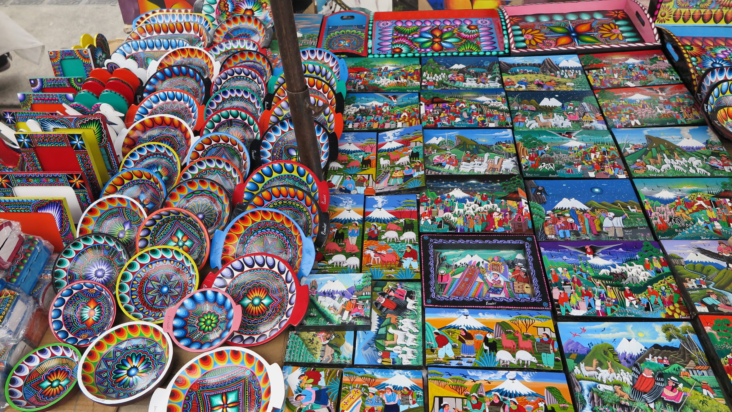 A vibrant display at Otavalo Market in Ecuador featuring hand-painted wooden bowls and Tigua-style paintings depicting Andean landscapes, volcanoes, and indigenous festivities.