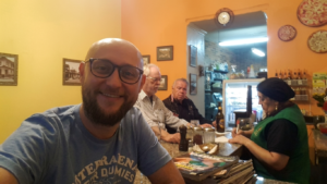 Ekvador'un Quito şehrindeki Café San Blas'ın tezgahında oturan; arka planda yerel müşteriler ve bir çalışanla birlikte gözlüklü ve sakallı erkek bir gezginin neşeli selfie karesi.