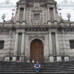 A traveler sitting on the stone steps leading up to the grand stone facade of the historic Iglesia de San Francisco in Quito, Ecuador.
