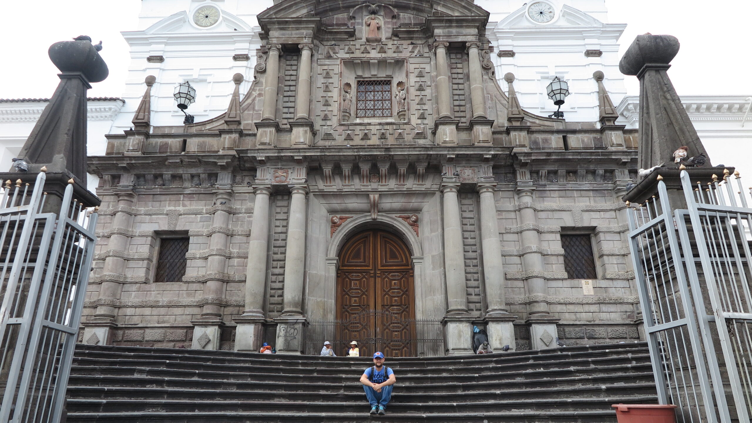 A traveler sitting on the stone steps leading up to the grand stone facade of the historic Iglesia de San Francisco in Quito, Ecuador.