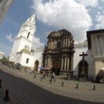 A dramatic wide-angle view of the historic dark stone facade of Iglesia Católica de El Sagrario next to a tall white bell tower on a cobblestone street in Quito, Ecuador.