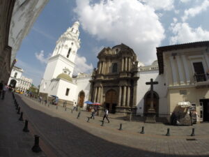 A dramatic wide-angle view of the historic dark stone facade of Iglesia Católica de El Sagrario next to a tall white bell tower on a cobblestone street in Quito, Ecuador.