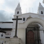 The white facade of Iglesia de El Carmen Alto and Museo del Carmen Alto in Quito, Ecuador, featuring twin bell towers, a stone cross, and an arched gateway.