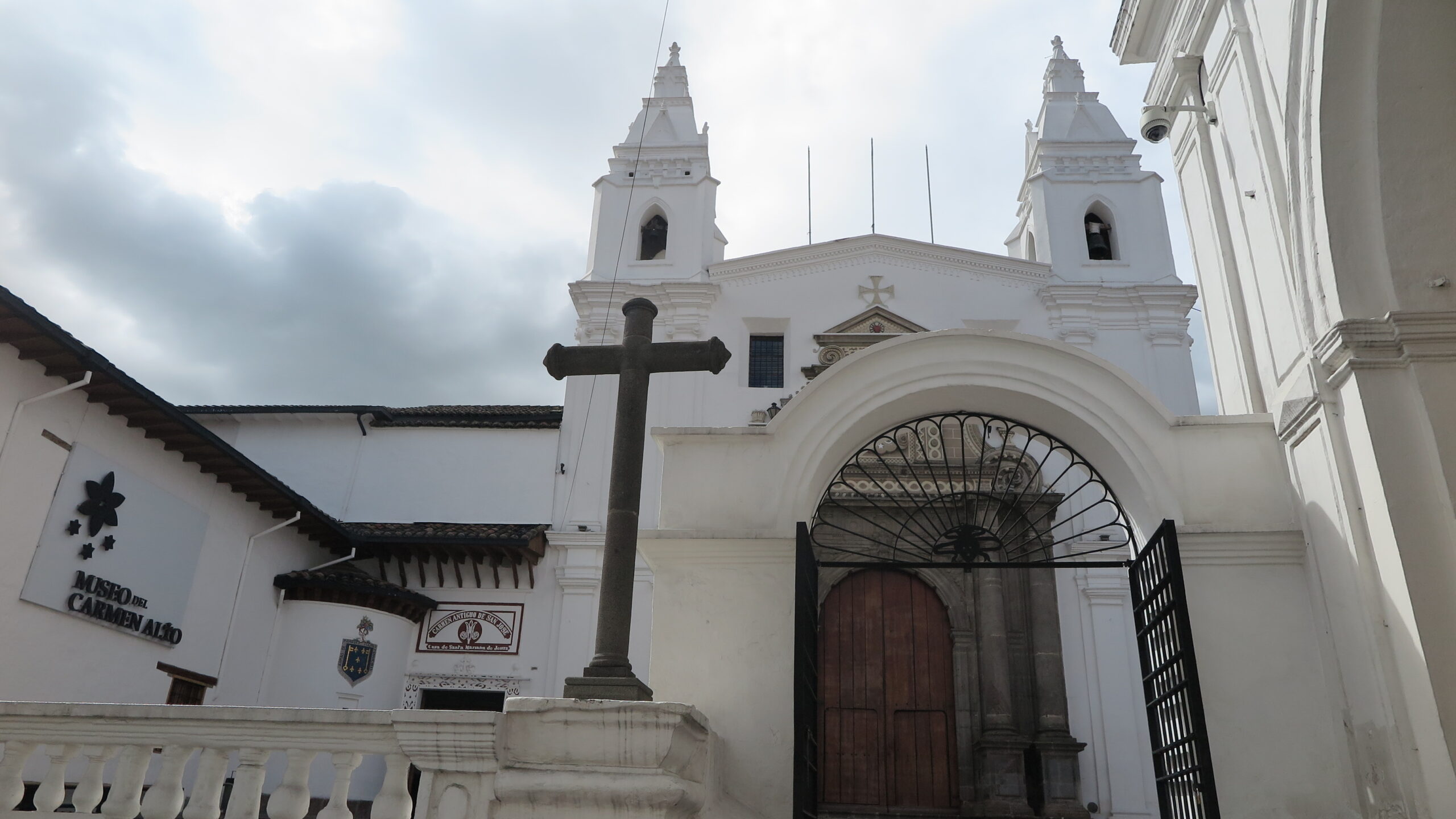 The white facade of Iglesia de El Carmen Alto and Museo del Carmen Alto in Quito, Ecuador, featuring twin bell towers, a stone cross, and an arched gateway.