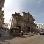 A wide-angle fish-eye shot of the ornate dark stone volcanic facade of Iglesia de la Compañía de Jesús on a historic street with pedestrians in Quito, Ecuador.