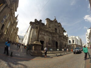 A wide-angle fish-eye shot of the ornate dark stone volcanic facade of Iglesia de la Compañía de Jesús on a historic street with pedestrians in Quito, Ecuador.