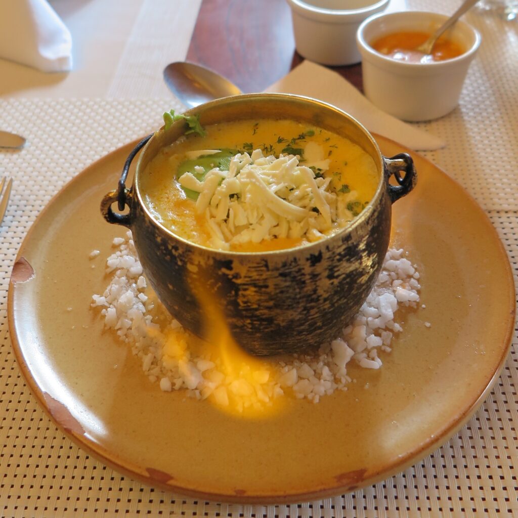 A close-up shot of a traditional Ecuadorian Locro Quiteño potato soup served in a rustic black cauldron pot topped with cheese and avocado, resting on a bed of coarse white salt.