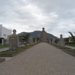A central stone walkway at Mitad del Mundo (Equator Line) lined with historical busts leading toward the iconic Equator Monument under an overcast sky in Quito, Ecuador.