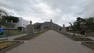 A central stone walkway at Mitad del Mundo (Equator Line) lined with historical busts leading toward the iconic Equator Monument under an overcast sky in Quito, Ecuador.