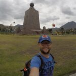 A close-up selfie of a male traveler with a backpack and a blue cap, smiling in front of the tall stone Equator Monument at Mitad del Mundo (Equator Line) in Quito, Ecuador.