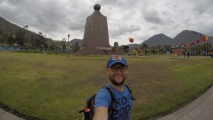 A close-up selfie of a male traveler with a backpack and a blue cap, smiling in front of the tall stone Equator Monument at Mitad del Mundo (Equator Line) in Quito, Ecuador.
