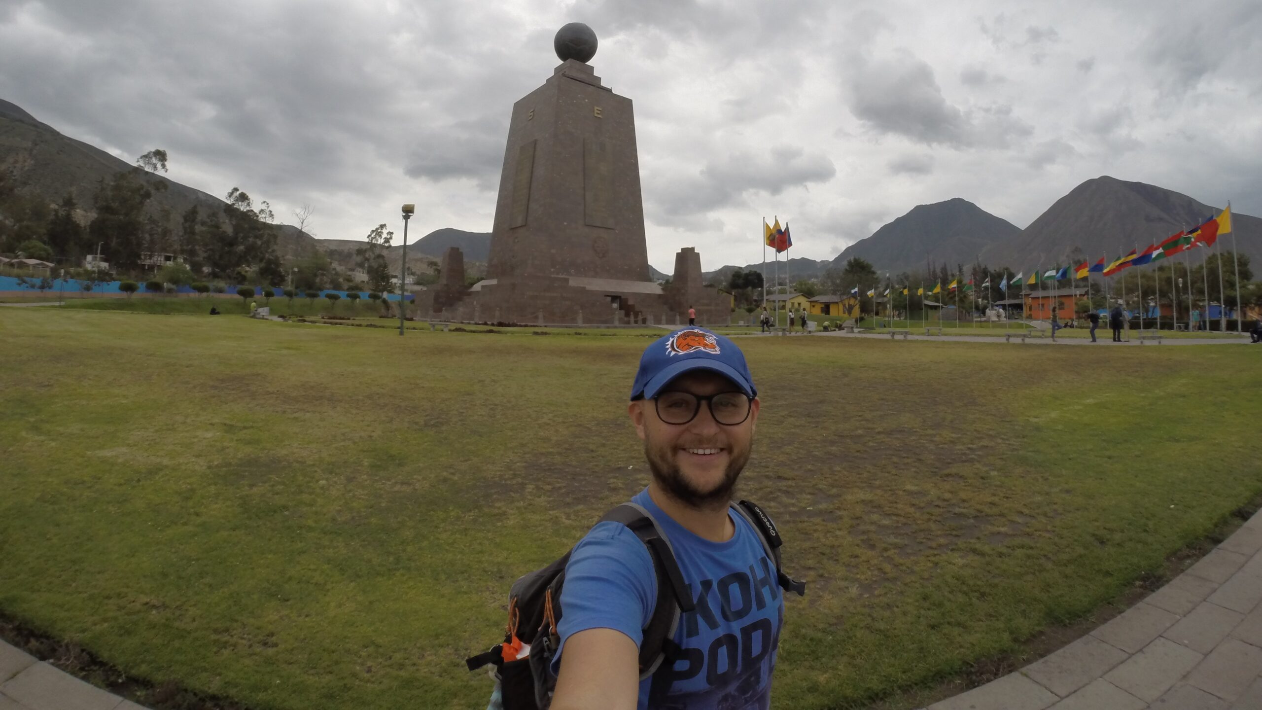 A close-up selfie of a male traveler with a backpack and a blue cap, smiling in front of the tall stone Equator Monument at Mitad del Mundo (Equator Line) in Quito, Ecuador.