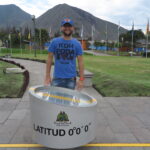 A male traveler in a blue t-shirt and blue cap standing behind a silver cylindrical sign reading "LATITUD 0°0'0"" along a yellow line at Mitad del Mundo (Equator Line) in Quito, Ecuador.