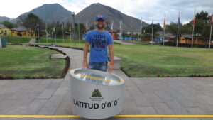 A male traveler in a blue t-shirt and blue cap standing behind a silver cylindrical sign reading "LATITUD 0°0'0"" along a yellow line at Mitad del Mundo (Equator Line) in Quito, Ecuador.