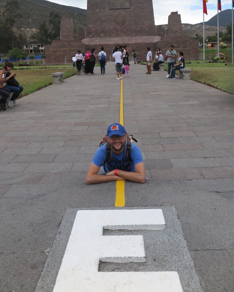 A male traveler in a blue t-shirt and cap lying face down along the yellow equator line in front of the stone monument at Mitad del Mundo (Equator Line) in Quito, Ecuador.