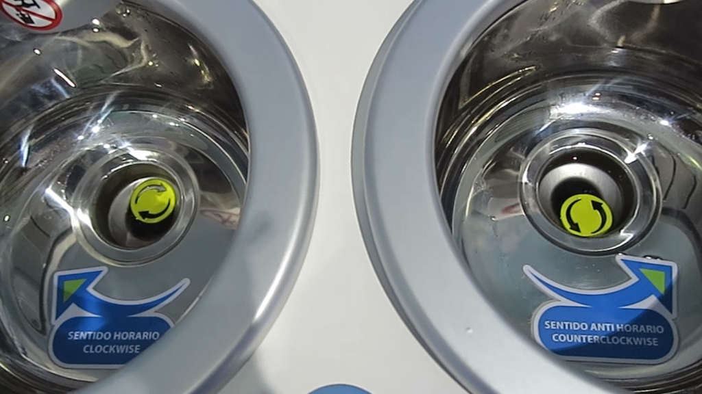 A close-up of two stainless steel sinks side by side at the Equator, one labeled "Clockwise" and the other "Counterclockwise" with directional arrows inside.