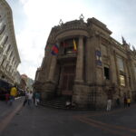 A wide-angle fish-eye shot of the grand stone neoclassical facade of the Museo de la Moneda featuring tall columns and flags in Quito, Ecuador.