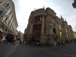 A wide-angle fish-eye shot of the grand stone neoclassical facade of the Museo de la Moneda featuring tall columns and flags in Quito, Ecuador.