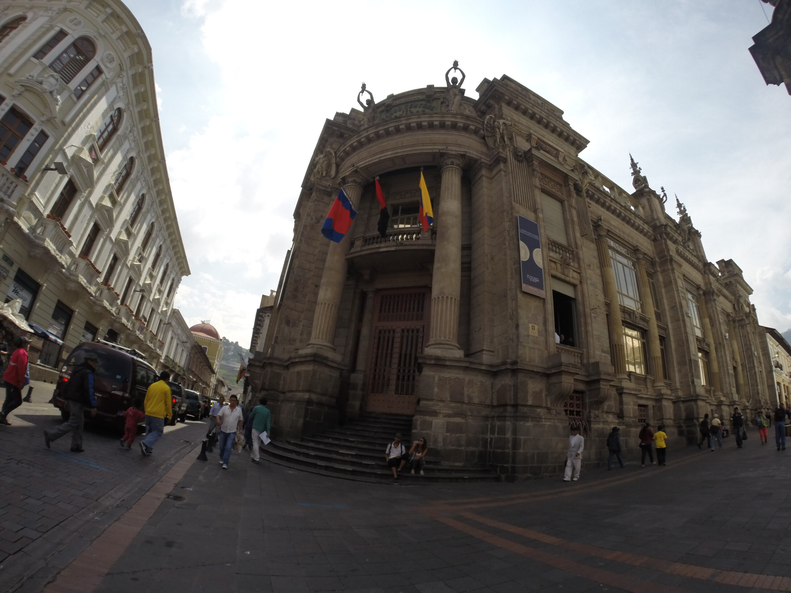 A wide-angle fish-eye shot of the grand stone neoclassical facade of the Museo de la Moneda featuring tall columns and flags in Quito, Ecuador.