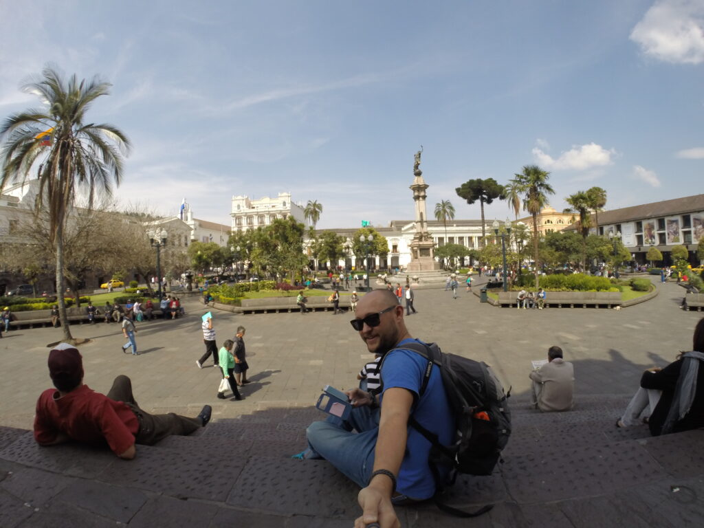 A wide-angle selfie of a traveler with a backpack sitting on the steps overlooking the historic Plaza Grande (Independence Square) in Quito, Ecuador, featuring the Independence Monument and colonial buildings in the background.